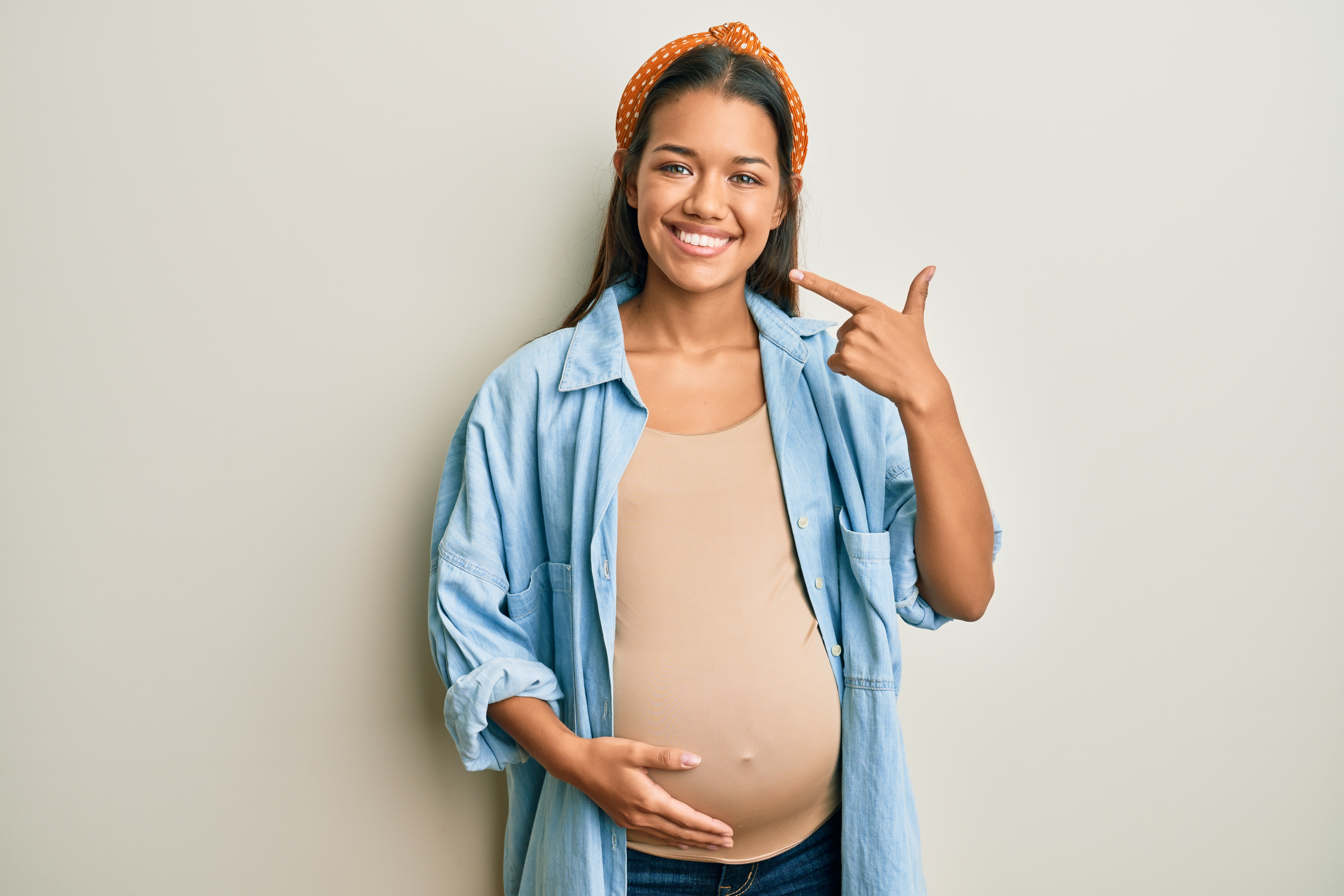 A pregnant woman holding her stomach and pointing at her smile.