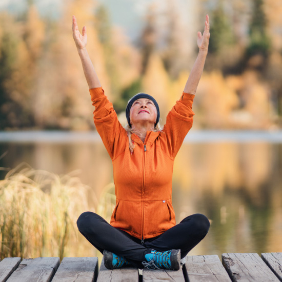 Person sitting cross-legged on a wooden dock, arms raised in the air, facing a serene lake surrounded by autumn trees